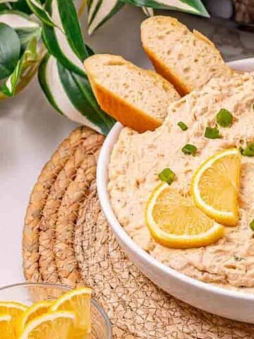 Creamy dip in a white bowl, garnished with lemon slices, green onions, and served with sliced baguette.