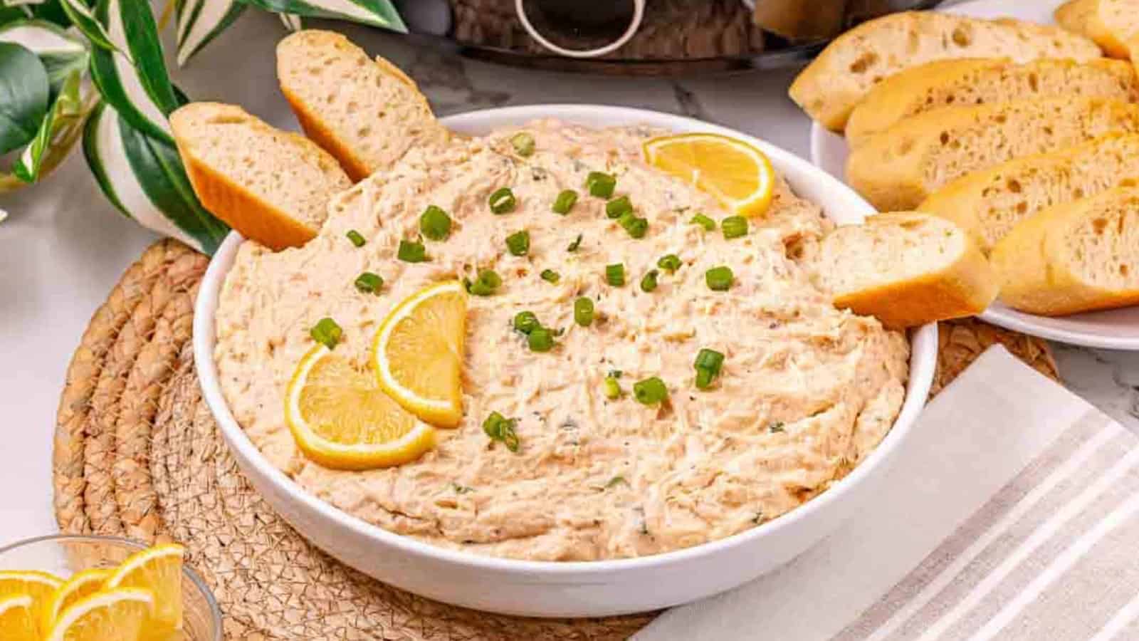 A bowl of creamy dip garnished with lemon slices and green onions, served with slices of baguette.
