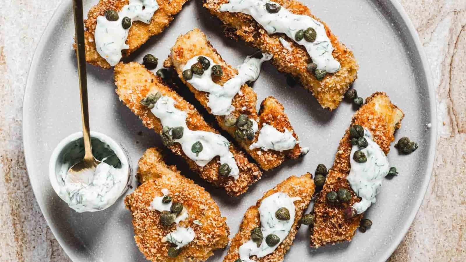Breaded fish sticks topped with creamy sauce and capers on a plate, with extra sauce in a small bowl.