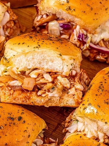 Close-up of pulled chicken sliders with coleslaw on golden, seasoned buns arranged on a wooden surface.