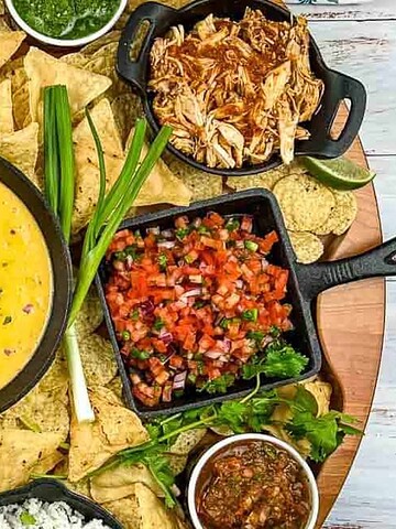A round platter with nacho chips, queso dip, shredded cheese, salsa, beans, chicken, rice, and various toppings.
