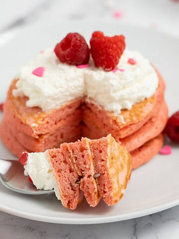 Stack of pink pancakes with whipped cream and raspberries, one bite cut and on a fork, on a white plate.