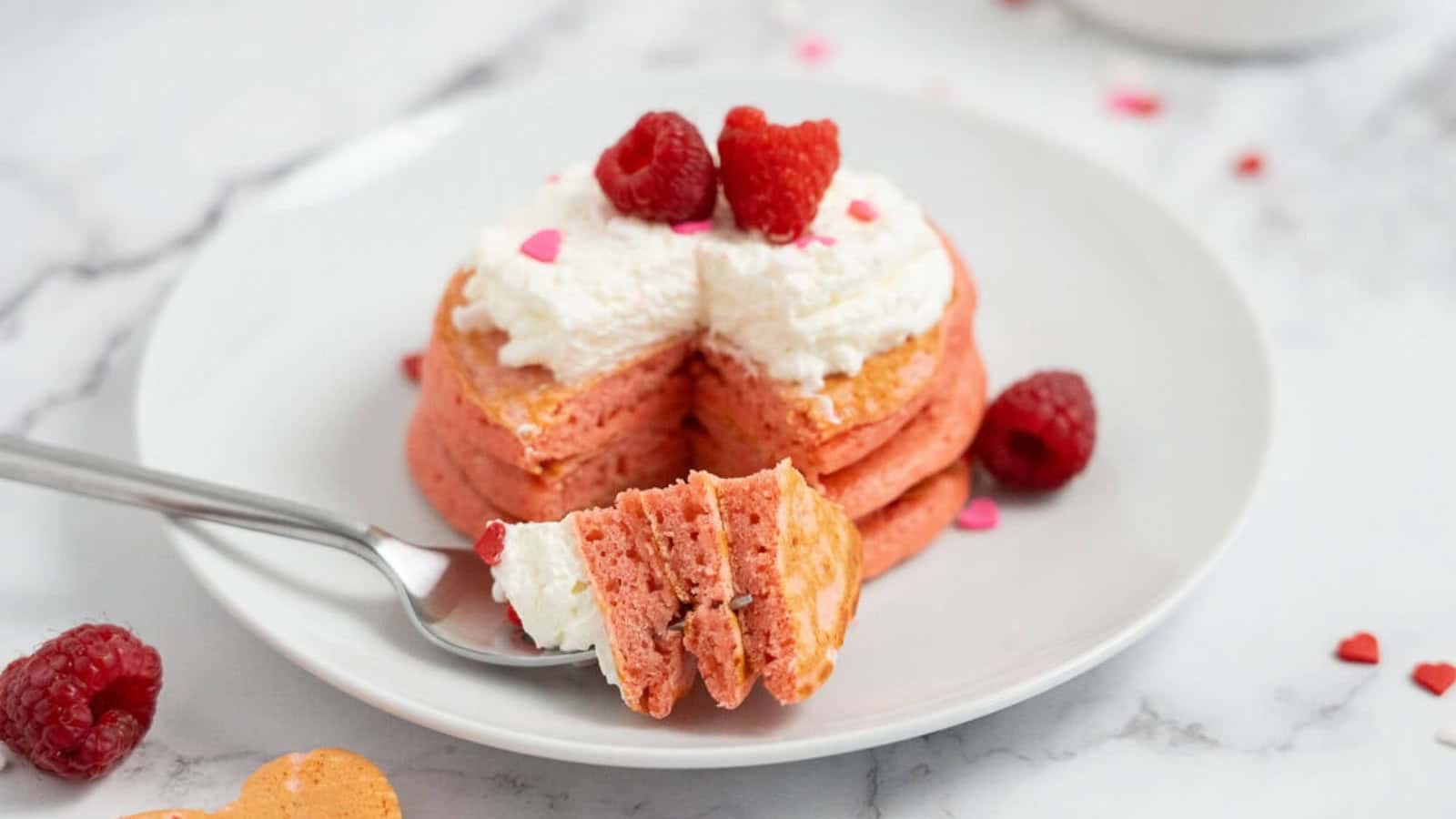 Stack of pink pancakes with whipped cream and raspberries, one bite cut and on a fork, on a white plate.