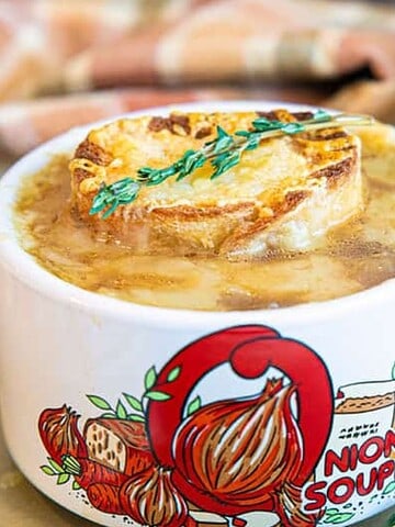 Two mugs of French onion soup with toasted bread and thyme, in white mugs decorated with onion illustrations.