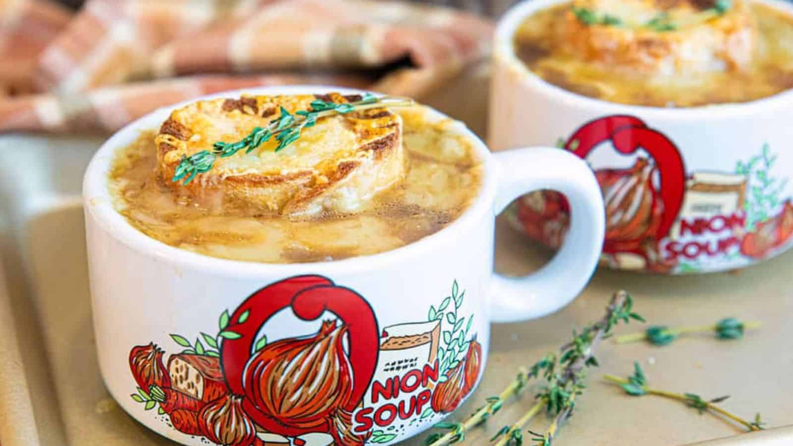 Two mugs of French onion soup with toasted bread and thyme, in white mugs decorated with onion illustrations.