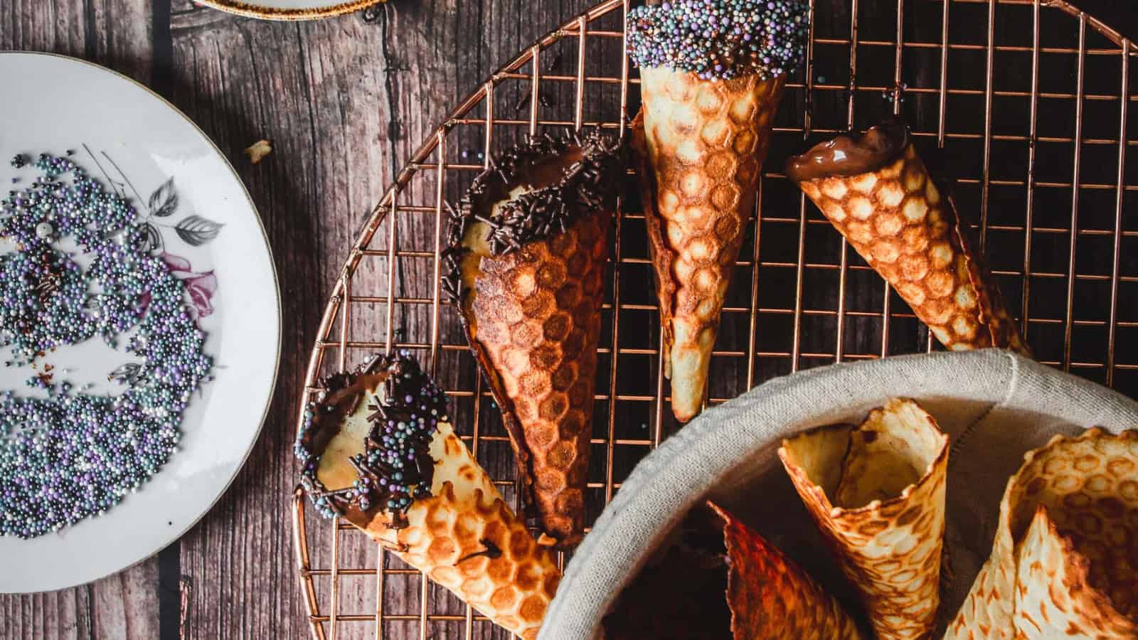 Waffle cones dipped in chocolate and sprinkles on a cooling rack, with a plate of sprinkles nearby.