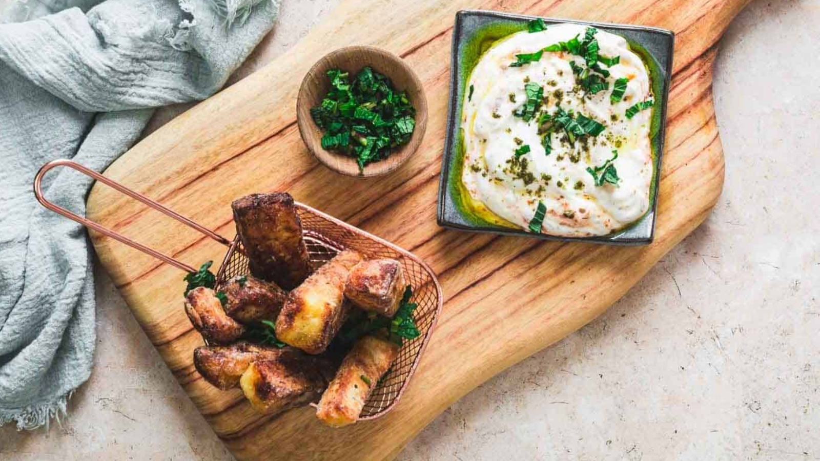 Fried halloumi sticks in a basket with creamy dip and chopped herbs on a wooden board.