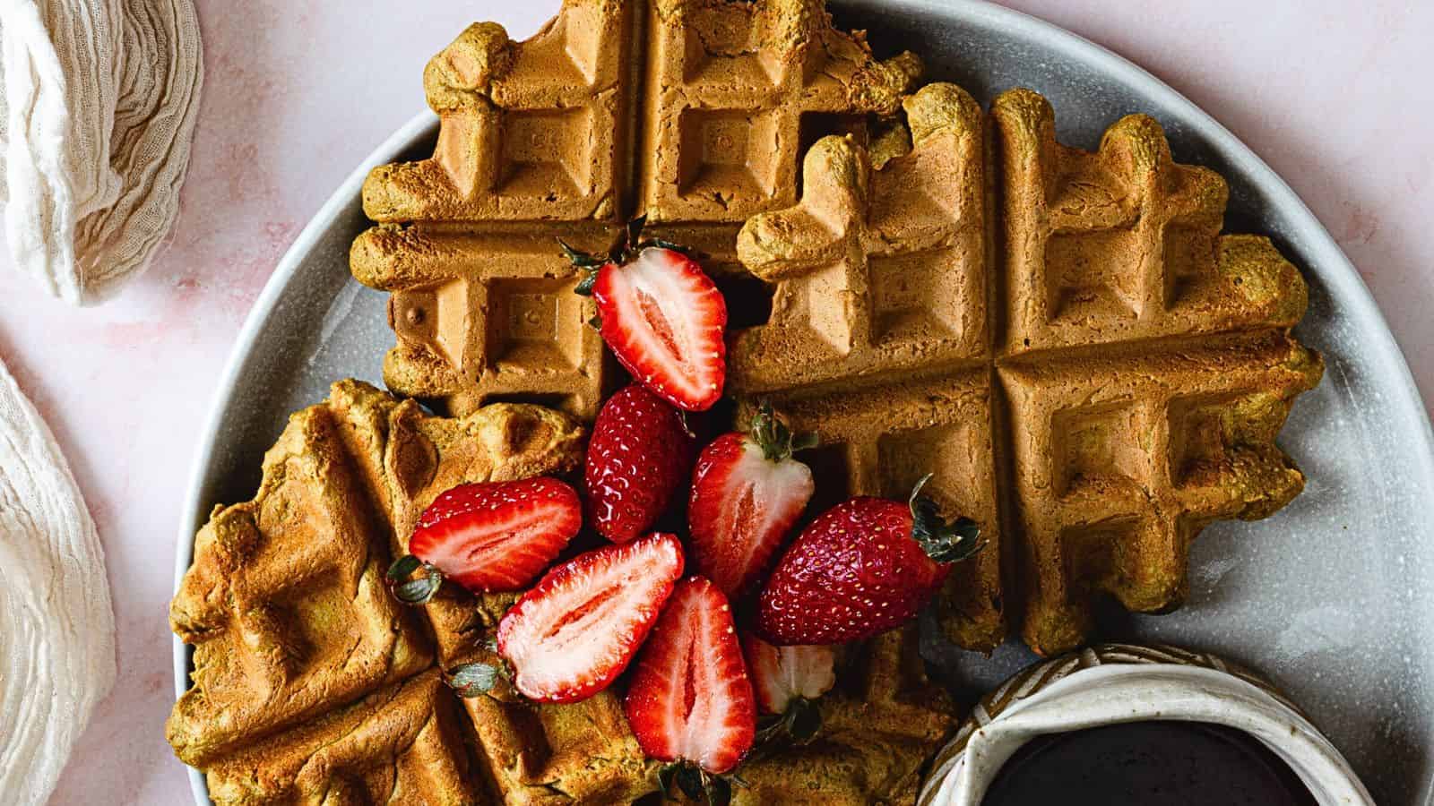A plate of four golden brown waffles topped with halved strawberries. A small jug of syrup is placed next to the waffles. The plate is on a light-colored surface, with two folded cloth napkins nearby.