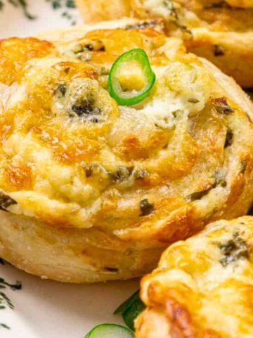 Close-up of golden cheese and herb pastry swirls topped with sliced green chili on a decorative plate.