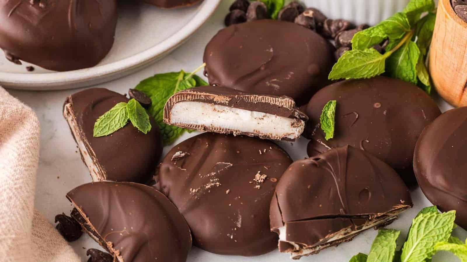 Chocolate-covered peppermint patties with mint leaves arranged on a surface. Some patties are whole, and others are cut to show the creamy filling.