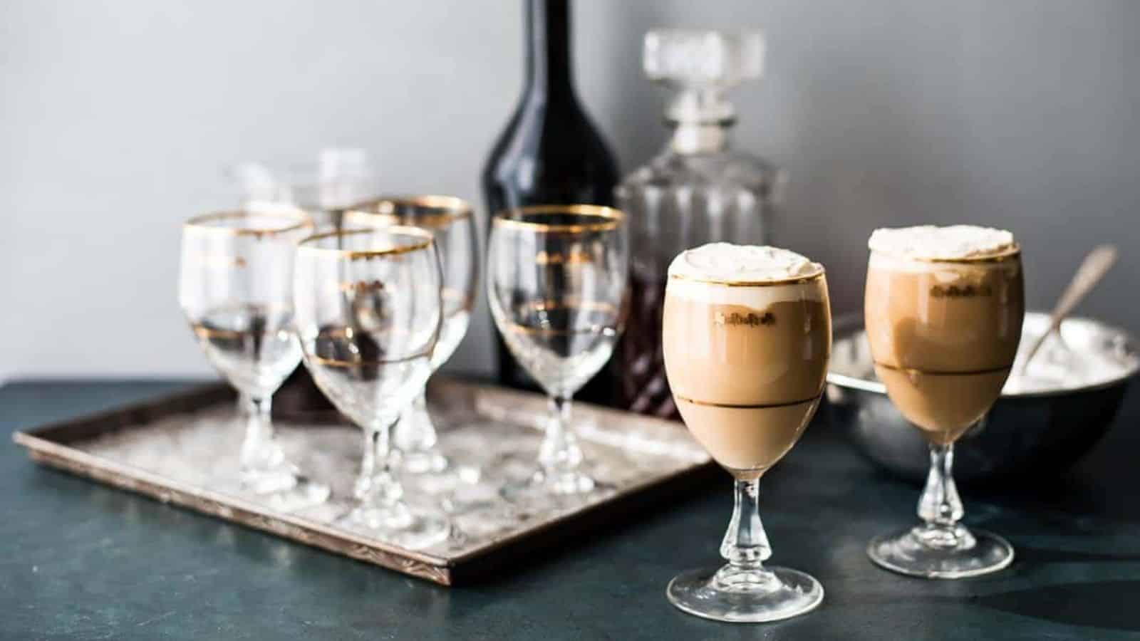 Two glasses of Irish Cream coffee in front of a tray of festive glass and a bar set up for making more.