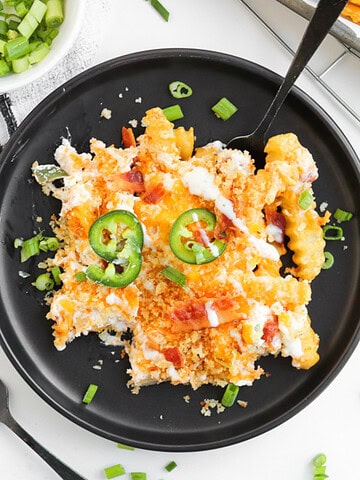 A serving of cheesy casserole topped with jalapeño slices and green onions on a black plate with a fork, inspired by the flavors of a jalapeno popper loaded cheese fries recipe.