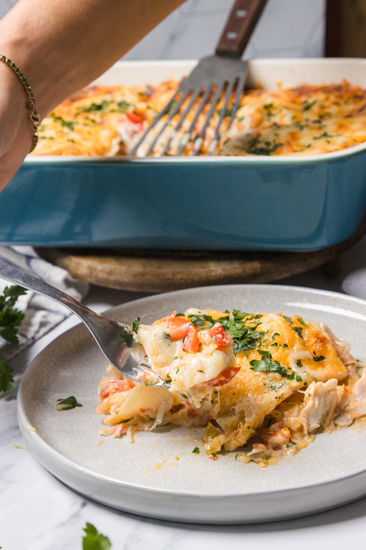 A hand serves cheesy King Ranch Chicken casserole onto a plate, garnished with herbs, with a blue dish in the background.