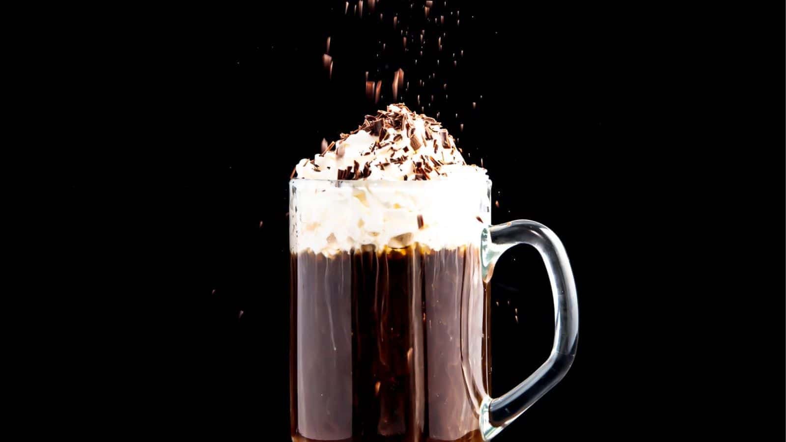A glass mug of hot chocolate with whipped cream and chocolate shavings against a black background.