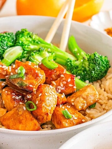A bowl of brown rice topped with broccoli, glazed tofu cubes, green onions, and sesame seeds.