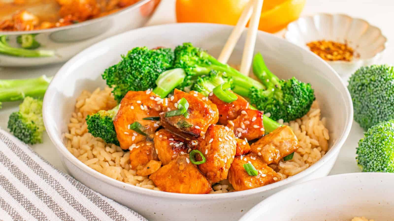 A bowl of brown rice topped with broccoli, glazed tofu cubes, green onions, and sesame seeds.