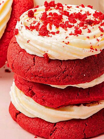 Three stacked red velvet cookies with cream cheese frosting and red crumbs on a pink surface.