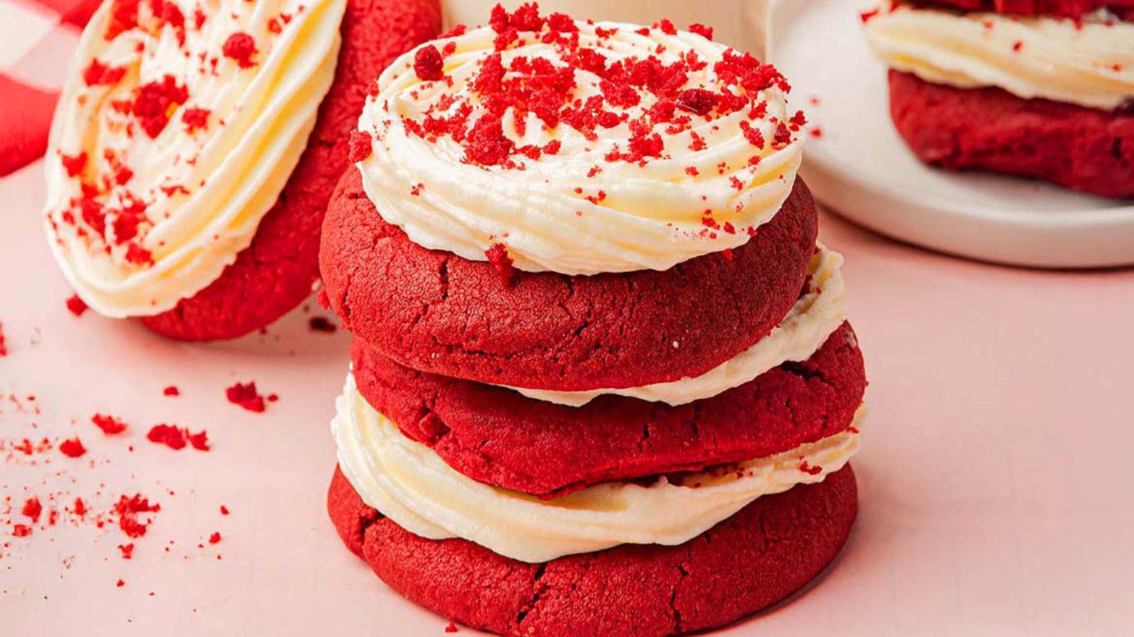Three red velvet cookies with white frosting are stacked on a pink surface. Crumbled red velvet pieces are sprinkled on top.