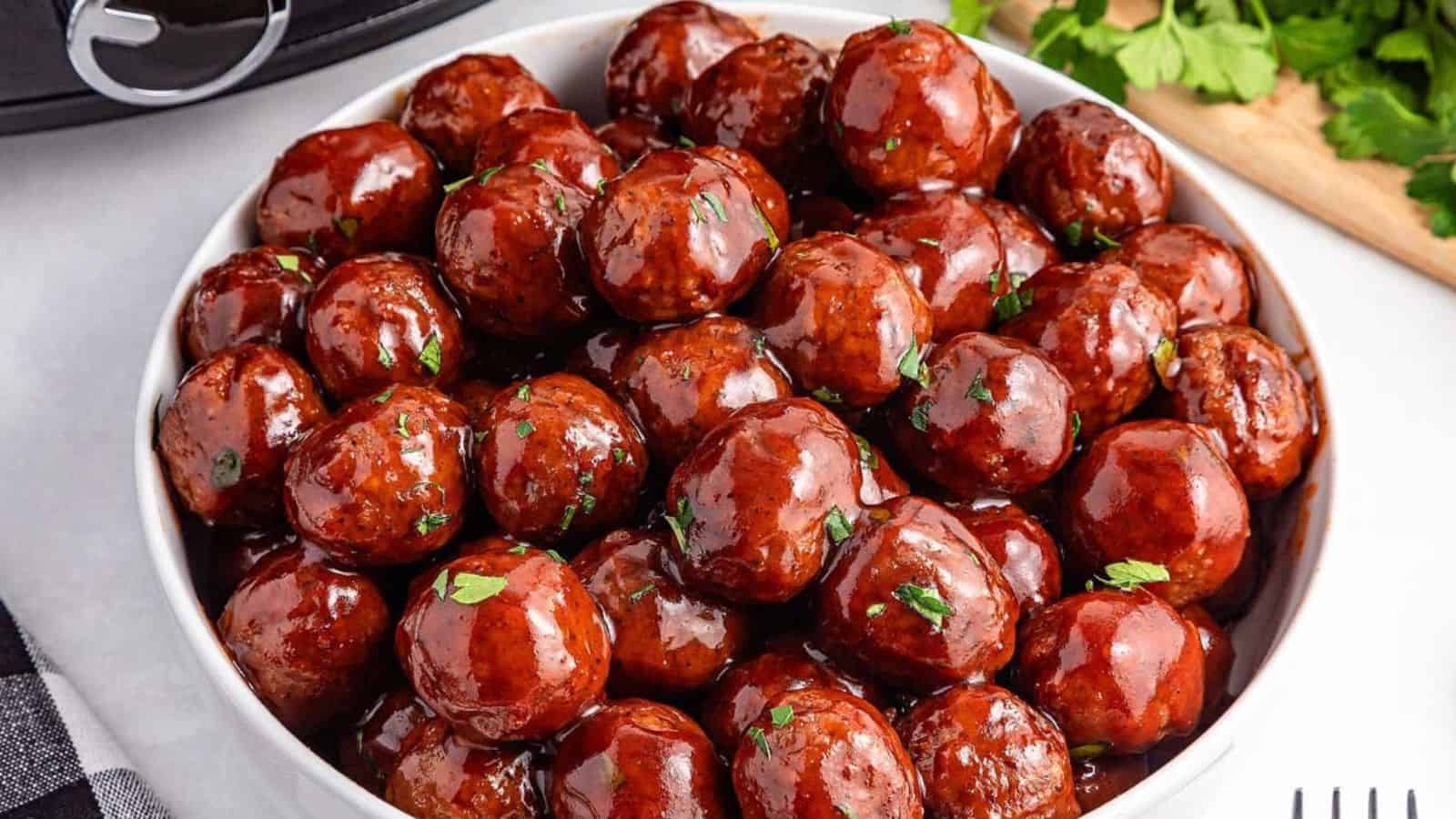 A bowl of shiny, glazed meatballs garnished with parsley on a white surface.