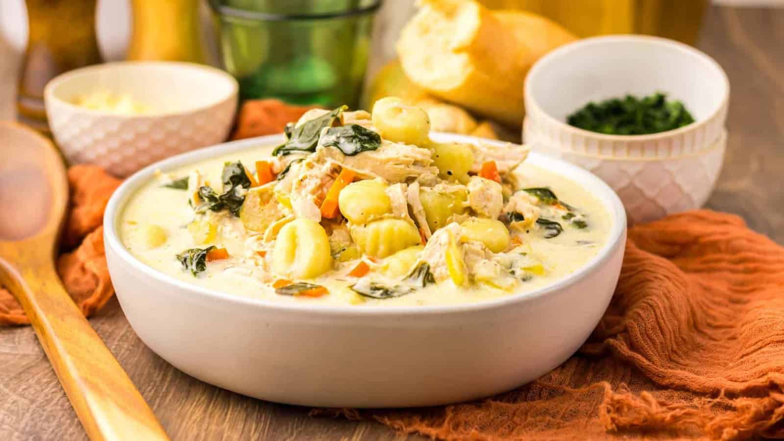 Creamy chicken gnocchi soup with vegetables served in a white bowl, bread and herbs on the side.