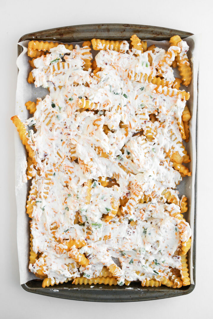 Crinkle-cut fries on a baking sheet topped with a creamy, white sauce, chopped herbs, and jalapeno popper loaded cheese for an irresistible fries recipe.