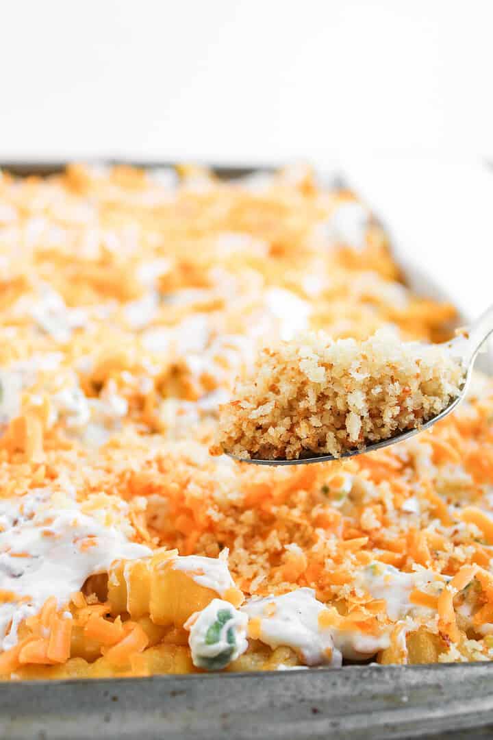A spoon holds a scoop of cheesy casserole&mdash;reminiscent of a jalapeno popper loaded cheese fries recipe&mdash;topped with crispy breadcrumbs above a baking dish of the same casserole.