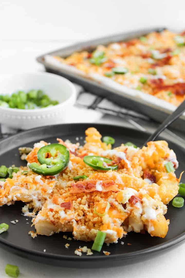 Plate of cheesy casserole topped with jalape&ntilde;o slices, bacon, and green onions&mdash;just like a jalapeno popper loaded cheese fries recipe&mdash;served hot with a baking tray in the background.
