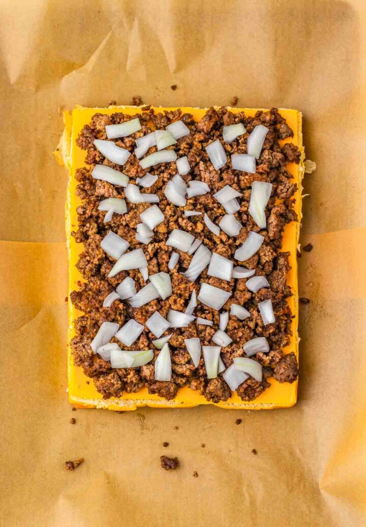 A rectangular pizza crust topped with cheese, ground meat, and chopped onions on parchment paper, inspired by the flavors of Big Mac Sliders for a unique homemade sliders twist.