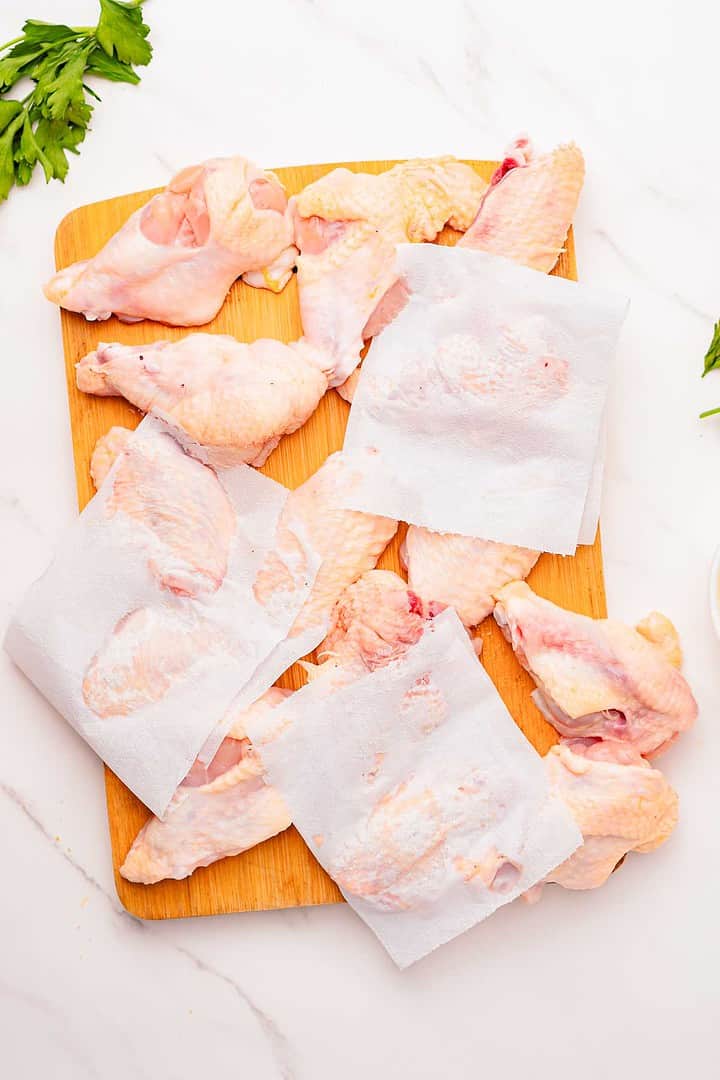 Raw chicken wings on a wooden board, some covered with paper towels; perfect for making garlic parmesan chicken wings. Parsley sits on the side.
