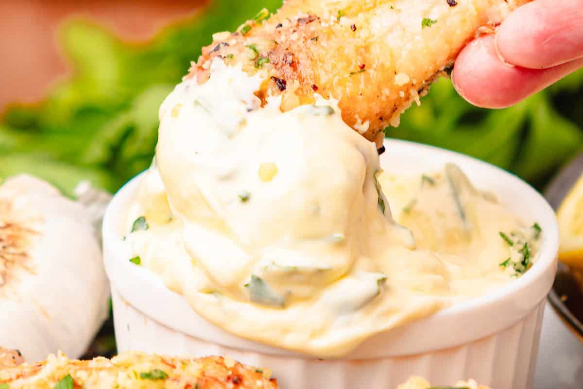 A hand dips a piece of breaded food, reminiscent of Garlic Parmesan Chicken Wings, into a creamy, herb-filled sauce in a small white bowl—perfect for enjoying air fryer creations.