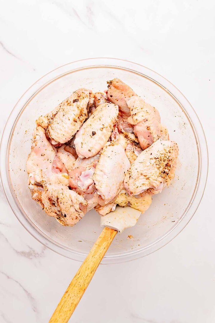 Raw chicken wings with seasoning in a glass bowl, being mixed with a spatula on a white marble surface—perfect for making Garlic Parmesan chicken wings in your air fryer.