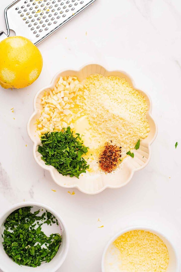 Scalloped bowl with chopped parsley, grated cheese, garlic, chili flakes, and a lemon nearby on a white surface—perfect for topping Air Fryer Garlic Parmesan Chicken Wings.