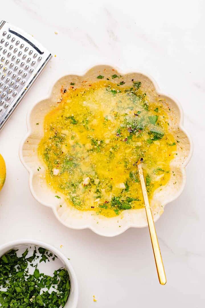 Bowl of yellow vinaigrette with herbs, a gold spoon, a grater, and chopped herbs on a white surface—perfect for drizzling over Garlic Parmesan chicken wings or using as an air fryer marinade.