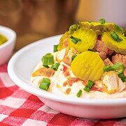 A bowl of creamy dip topped with sliced pickles, bacon bits, and chopped green onions on a red checkered cloth.