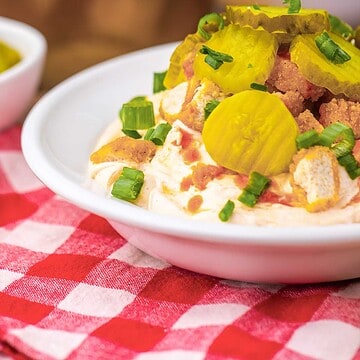A bowl of creamy dip topped with sliced pickles, bacon bits, and chopped green onions on a red checkered cloth.