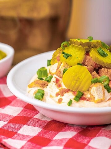 A bowl of creamy dip topped with sliced pickles, bacon bits, and chopped green onions on a red checkered cloth.