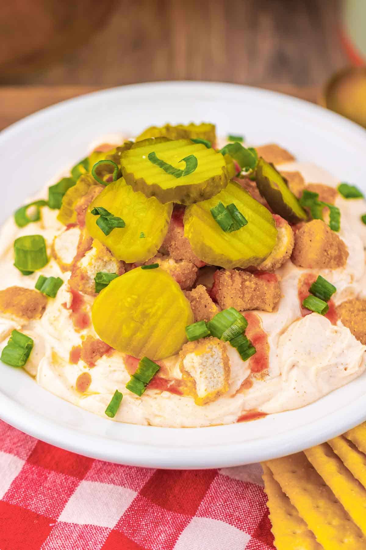 Bowl of creamy Chicken Dip topped with pickles, crushed crackers, green onions, and a spicy red sauce, served on a red plaid cloth&mdash;perfect for fans of Nashville Hot Chicken flavor.