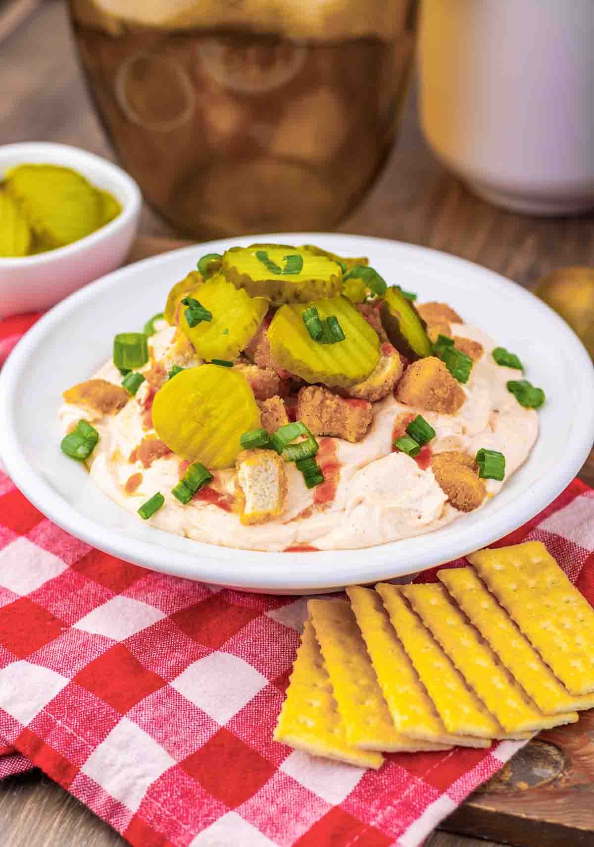 Creamy chicken dip inspired by Nashville Hot Chicken, topped with pickles, croutons, and green onions, served with crackers on a red checkered cloth.