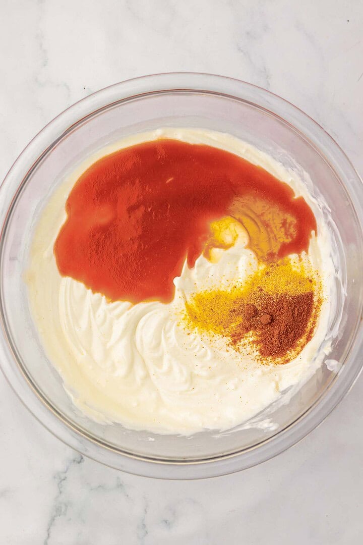 A glass bowl with white cream, red sauce, and yellow and brown spices on a marble surface&mdash;perfect for making a flavorful Nashville Hot Chicken dip.