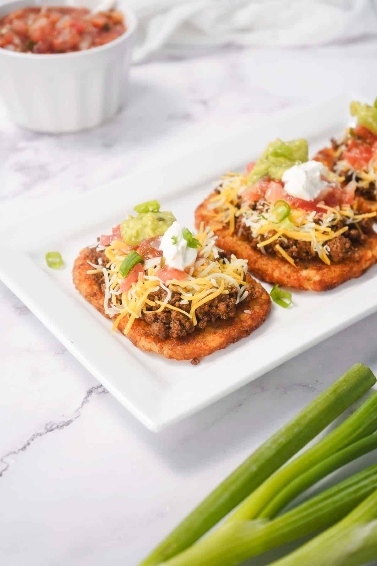 Two tostadas topped with savory ground beef, cheese, guacamole, sour cream, and tomatoes on a white plate—a delicious spin on the classic Beef Taco recipe.