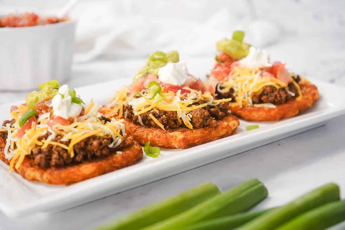Three loaded beef taco stacks with ground meat, cheese, hash brown, sour cream, and toppings are served on a white rectangular plate—a crave-worthy recipe for any taco lover.
