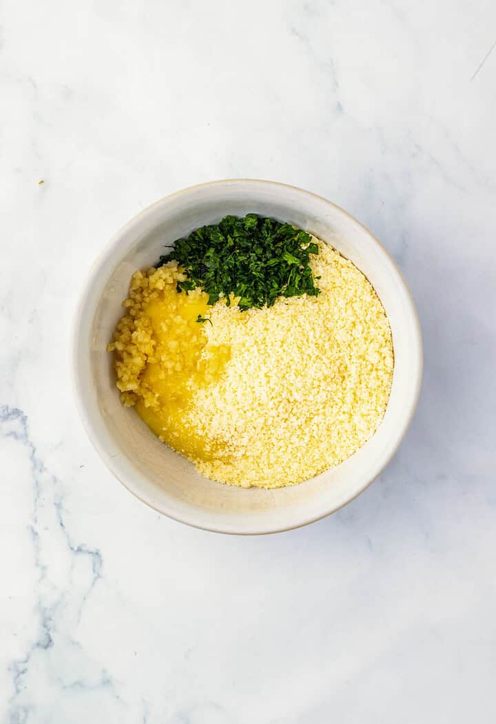 A bowl with grated cheese, minced garlic, chopped parsley, and melted butter on a white marble surface&mdash;perfect for preparing Parmesan chicken or adding flavor to your next sheet pan dinner.