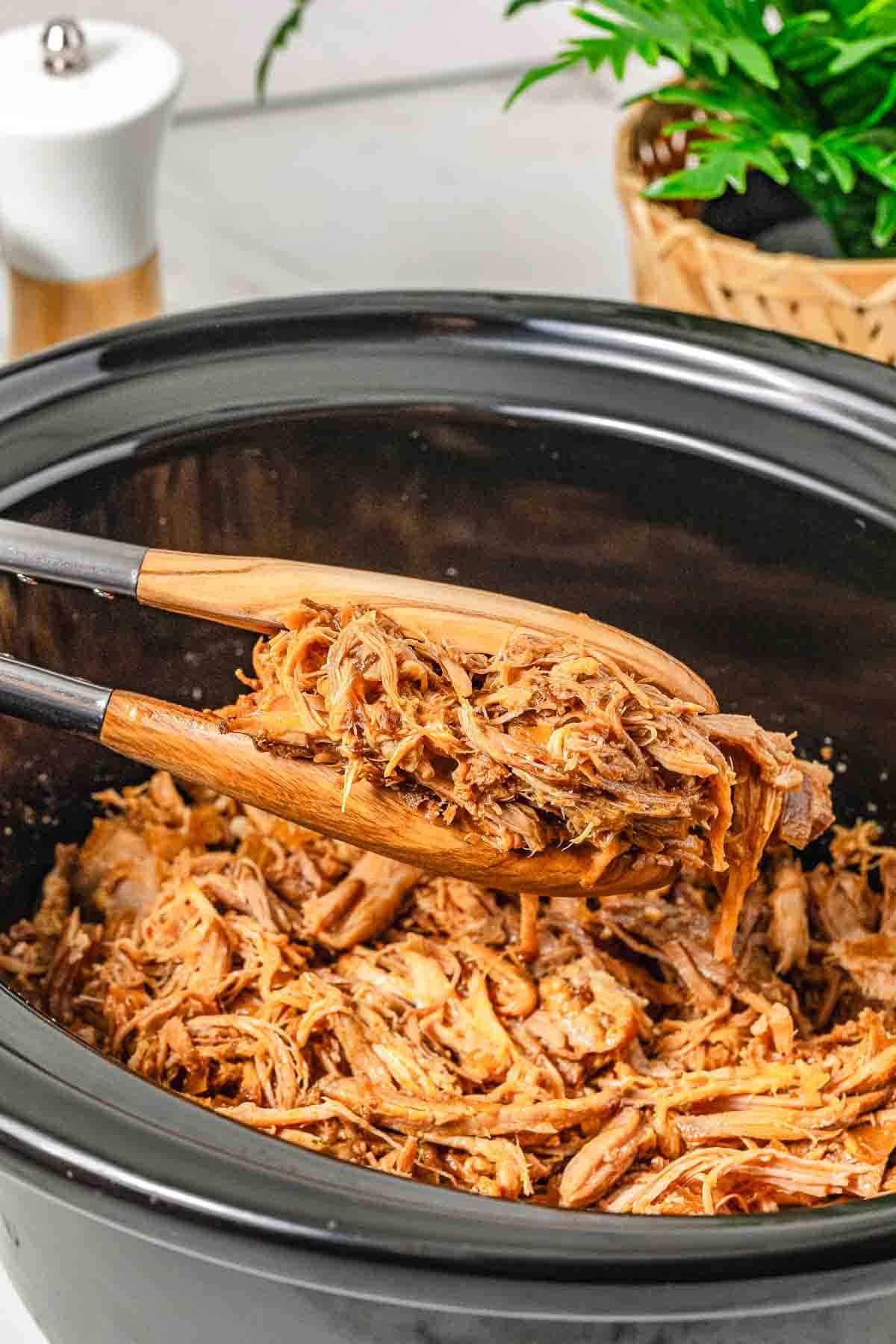 Tongs lifting tender BBQ pulled pork from a black slow cooker on a kitchen counter, ready to be piled onto a pulled pork sandwich.