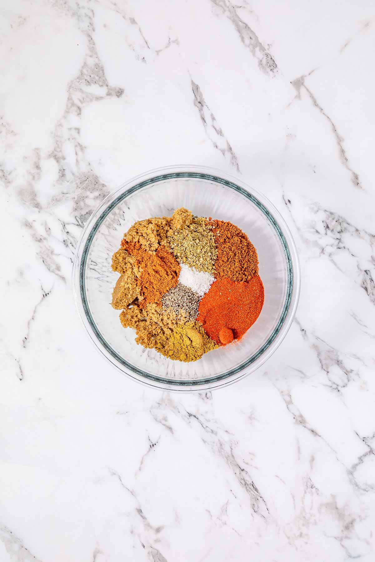 A glass bowl filled with various colorful ground spices on a white marble surface, perfect for seasoning your next BBQ Pulled Pork or Crockpot sandwich recipe.