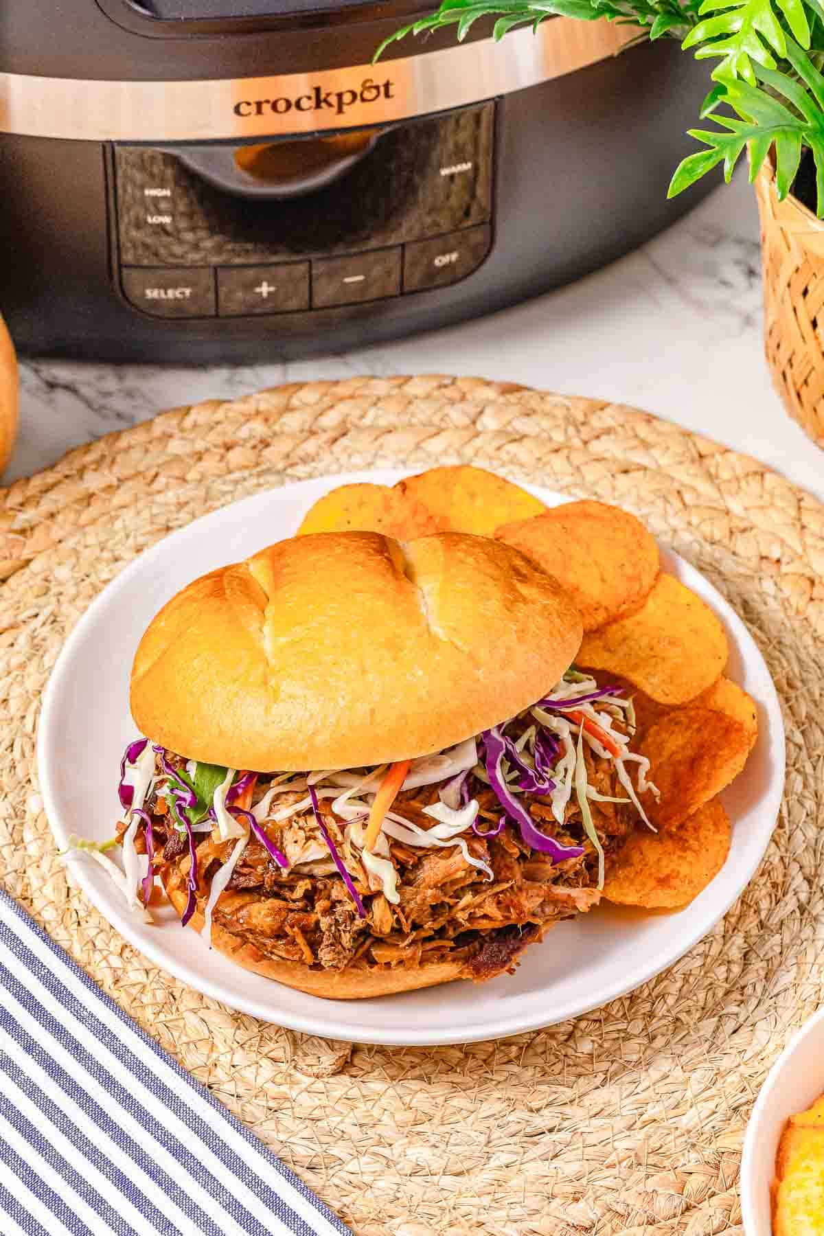 Pulled pork sandwich with coleslaw and chips on a white plate, showcasing a tasty BBQ Pulled Pork Sandwich Recipe; Crockpot slow cooker featured in the background.