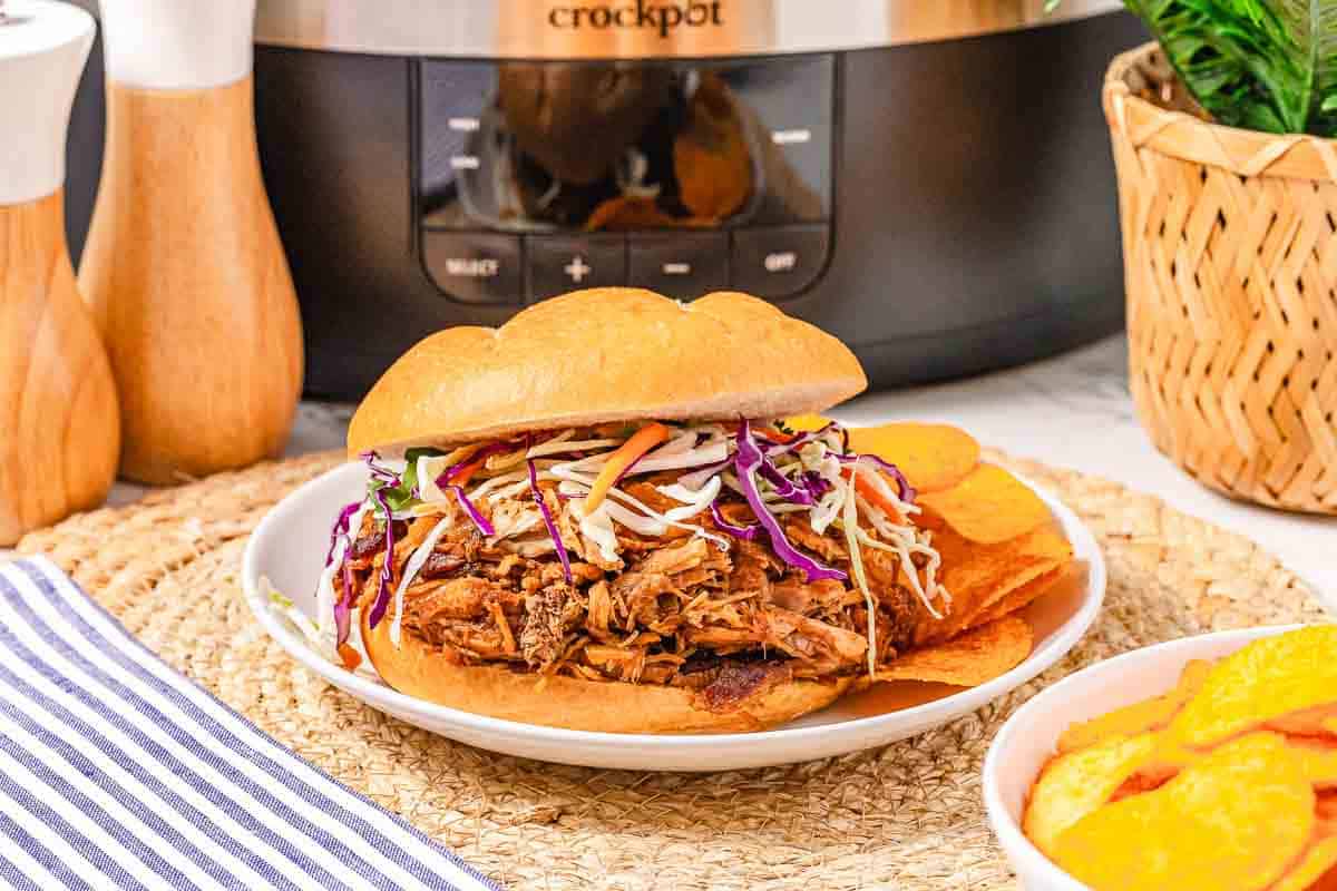 BBQ Pulled Pork sandwich with slaw on a plate, next to potato chips and a Crockpot in the background.