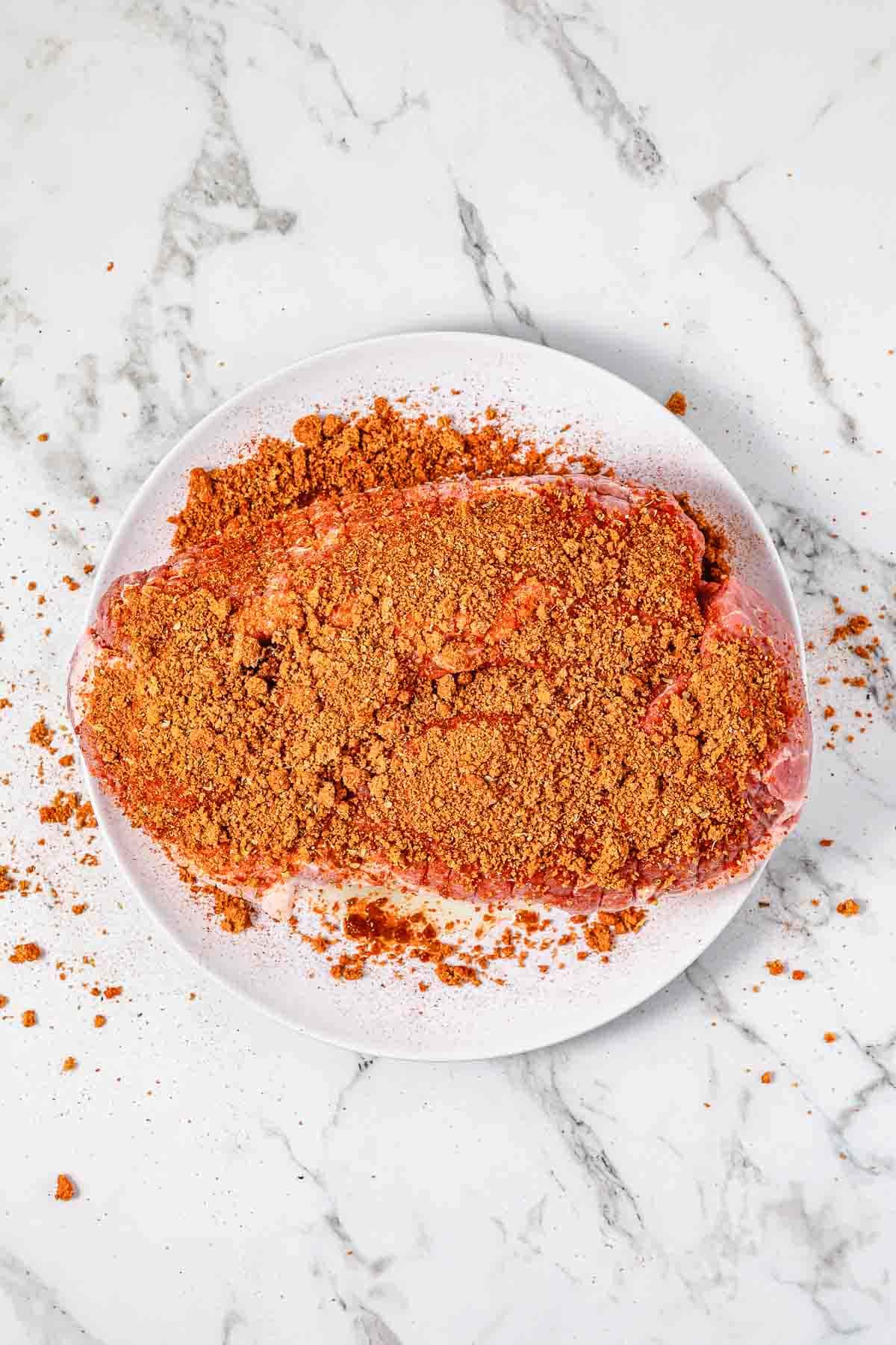 Raw steak on a white plate coated with a reddish-brown spice rub, perfect for transforming into a savory BBQ pulled pork or adding bold flavor to your favorite sandwich recipe. Set on a marble surface.