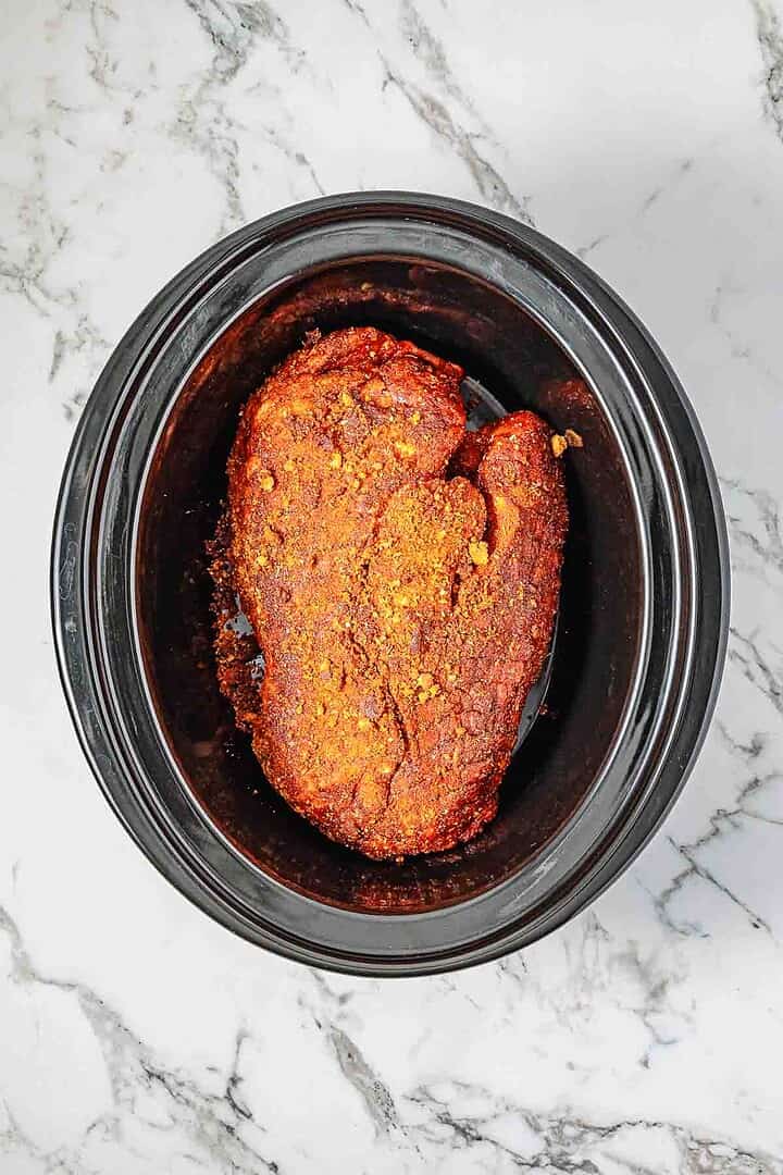 Seasoned pork shoulder in a black Crockpot on a white marble counter, ready to be cooked for the perfect BBQ Pulled Pork sandwich recipe.