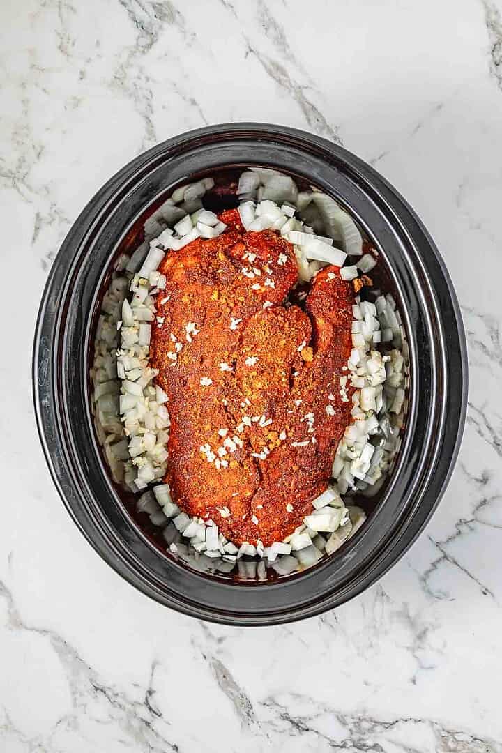 Raw seasoned meat and chopped onions sit in a black Crockpot on a white marble countertop—perfect for your next BBQ Pulled Pork sandwich recipe.