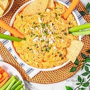 A bowl of creamy Buffalo Chicken Dip made in the crockpot, topped with chives and cheese, surrounded by celery, carrots, and tortilla chips.